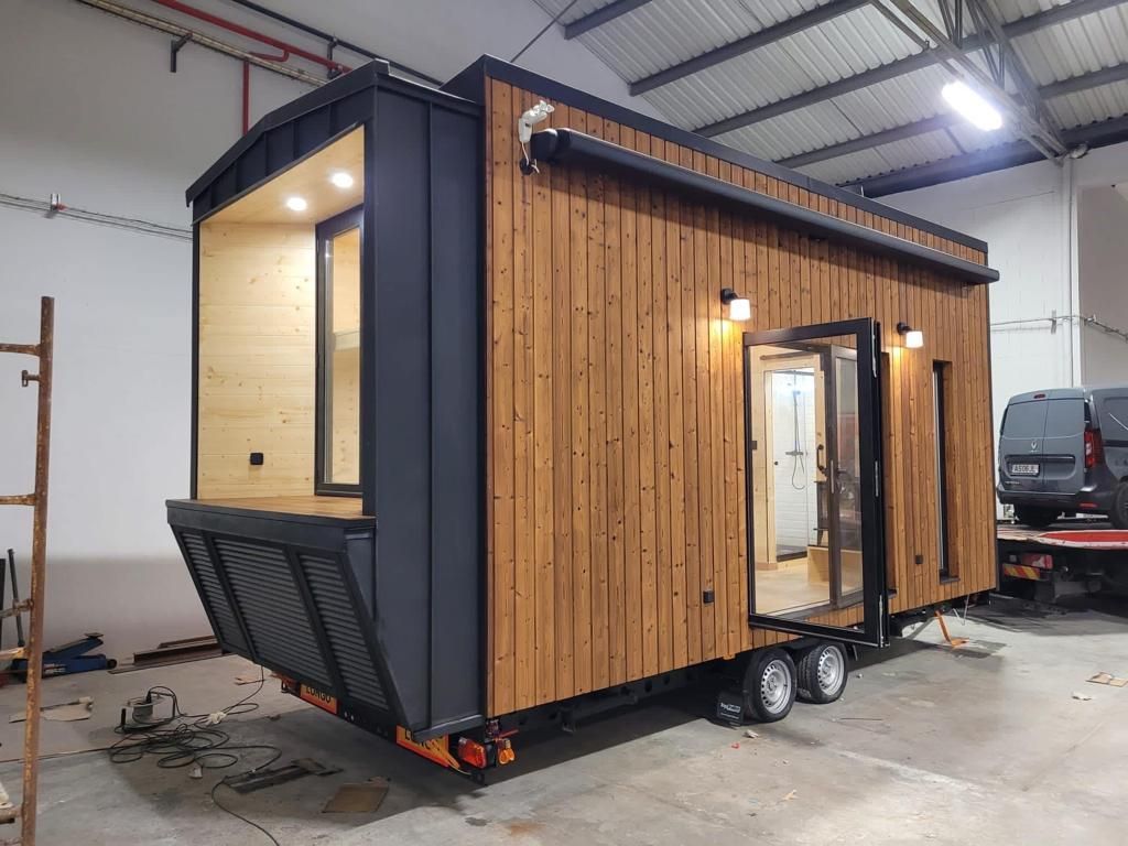 A tiny house on wheels with wooden siding, black accents, and an open door, parked inside a workshop.