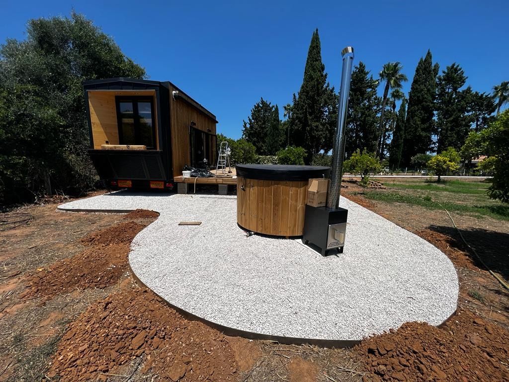 Tiny house with wooden hot tub on a gravel patio outdoors in a sunny setting.