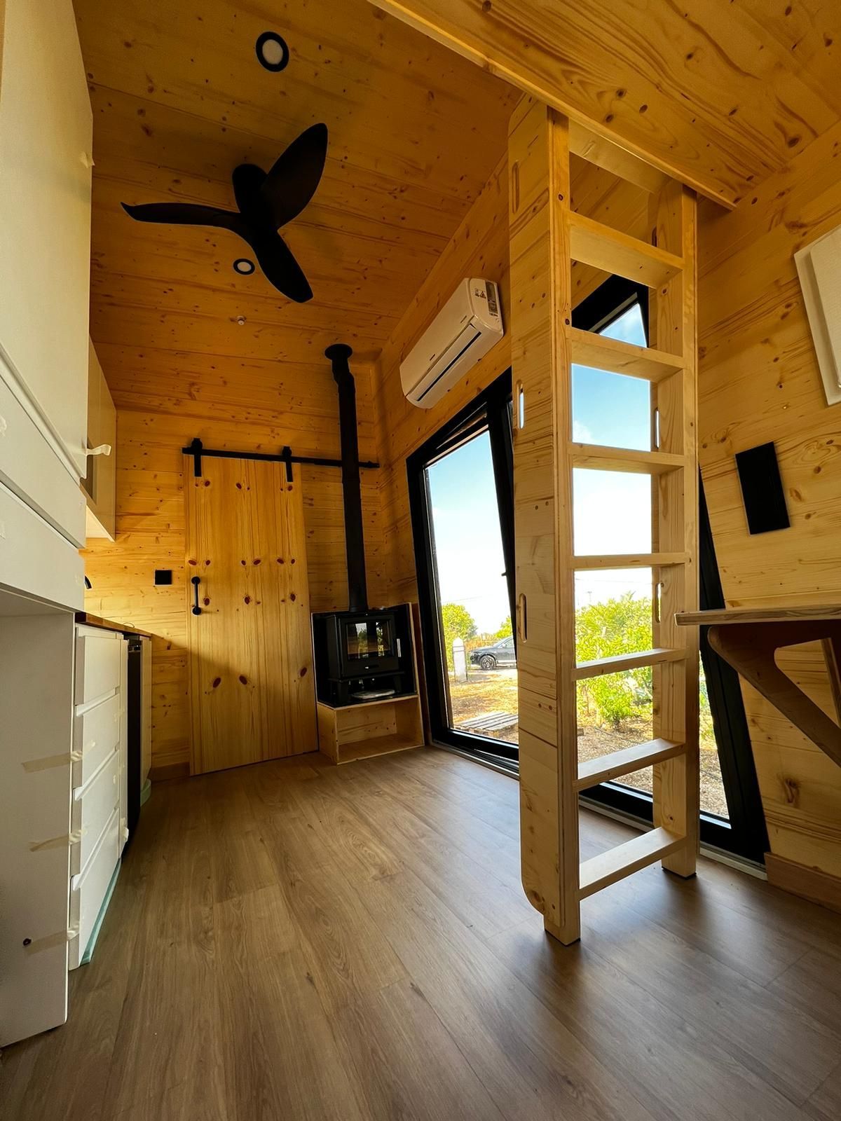 Wooden tiny home interior with ladder to loft, fireplace, and large window.
