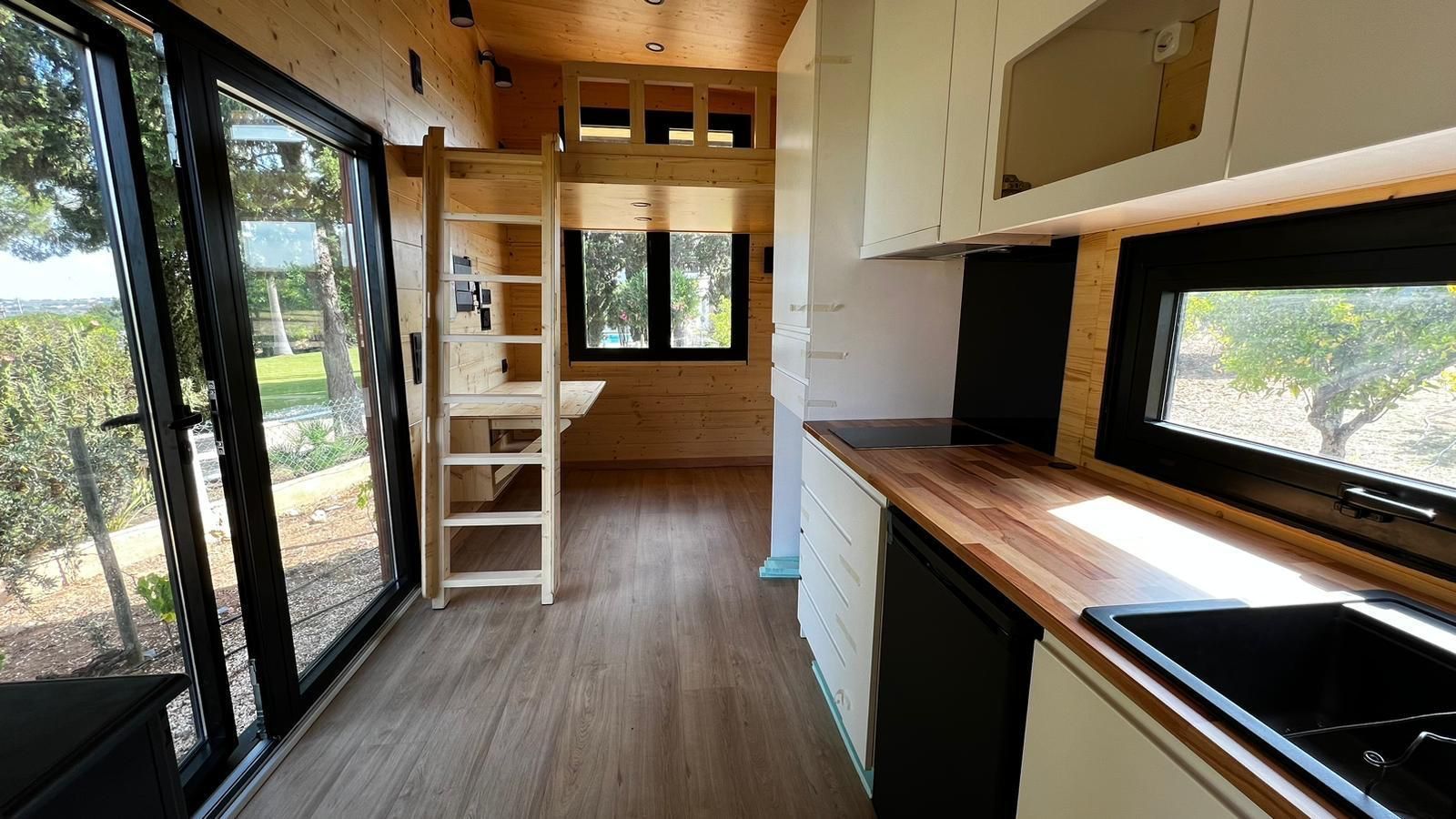 Interior of a tiny home with kitchen, desk, ladder to loft, and large windows overlooking greenery.