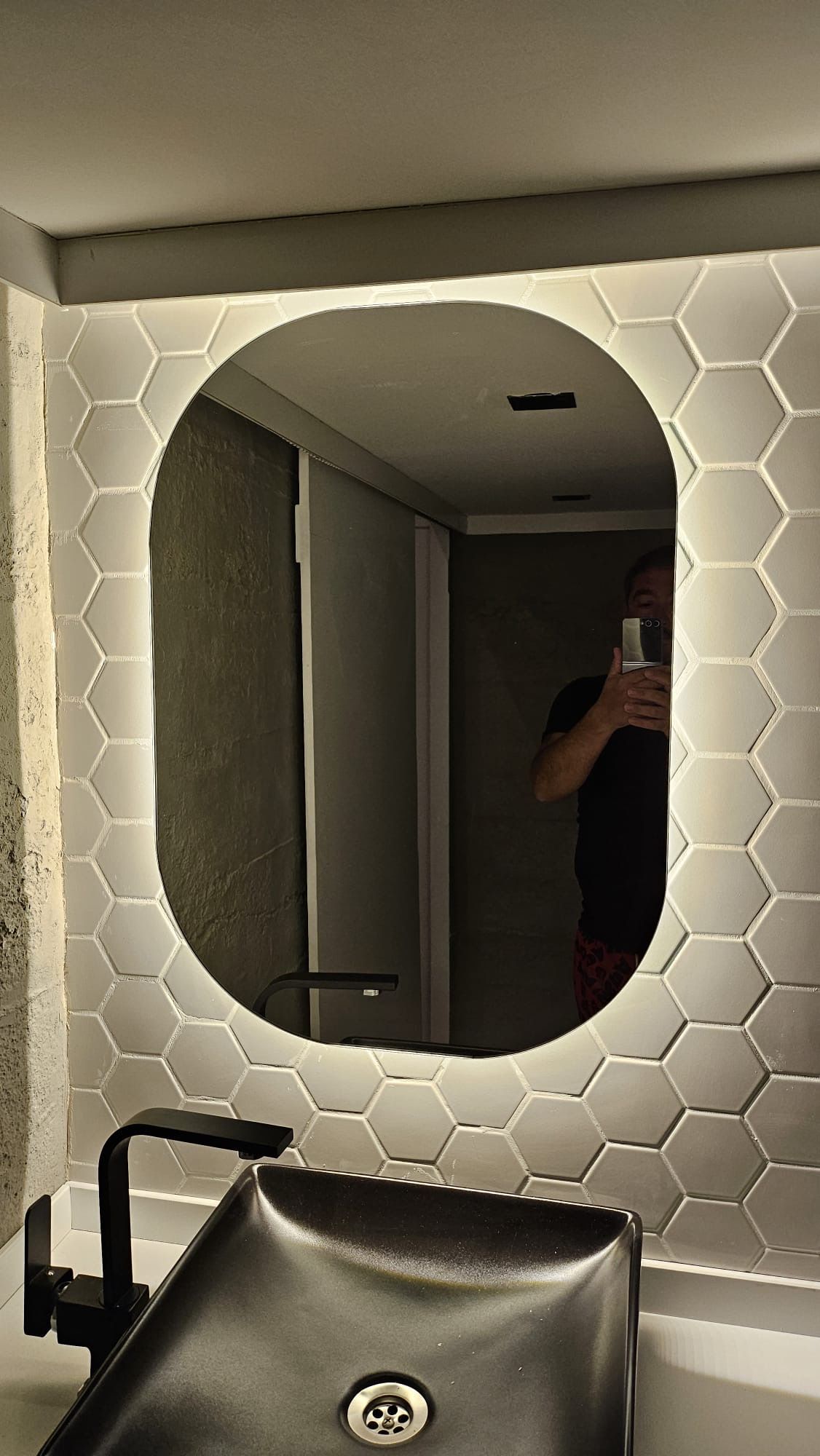 Oval mirror with LED lighting, set into a hexagon-tiled wall, above a black sink and faucet.