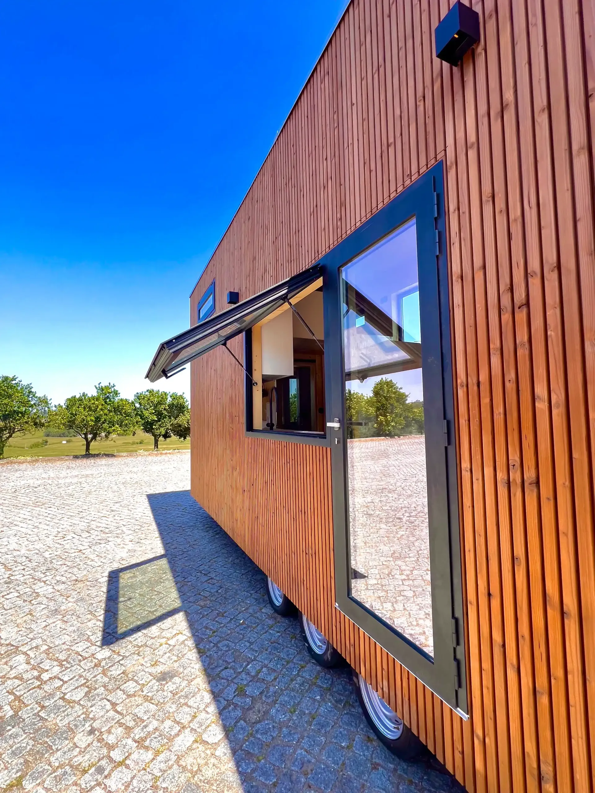 Tiny house with open window-door, brown wood siding, black trim, blue sky, parked on gravel.