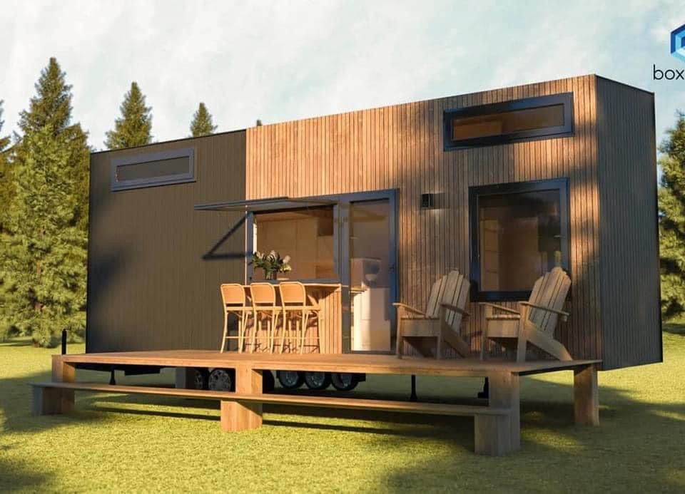 Tiny wooden house with deck, bar stools, Adirondack chairs, and two windows set in a grassy outdoor space.