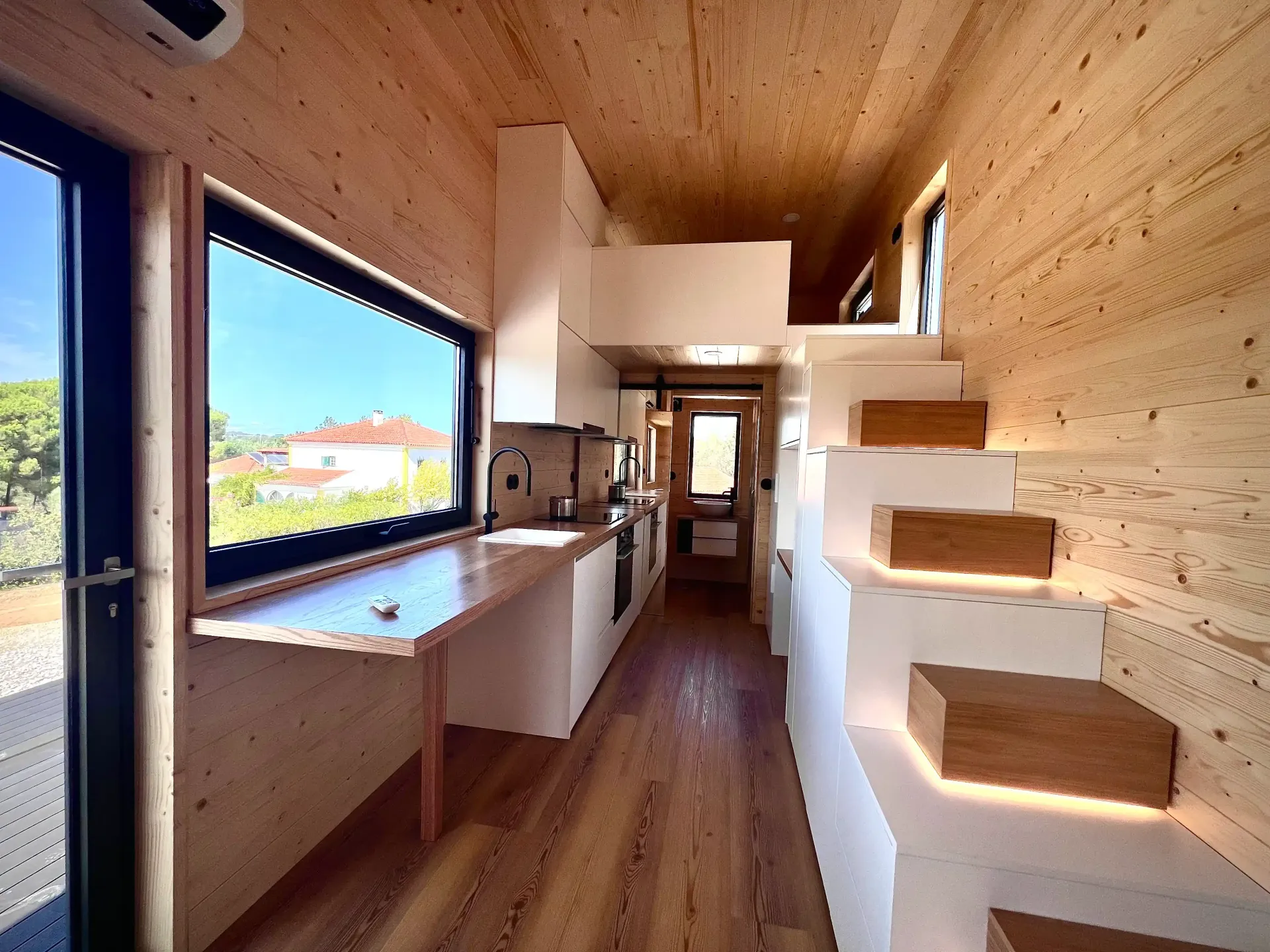 Interior of a tiny home with wooden walls, a window, stairs to a loft, and a kitchen area.