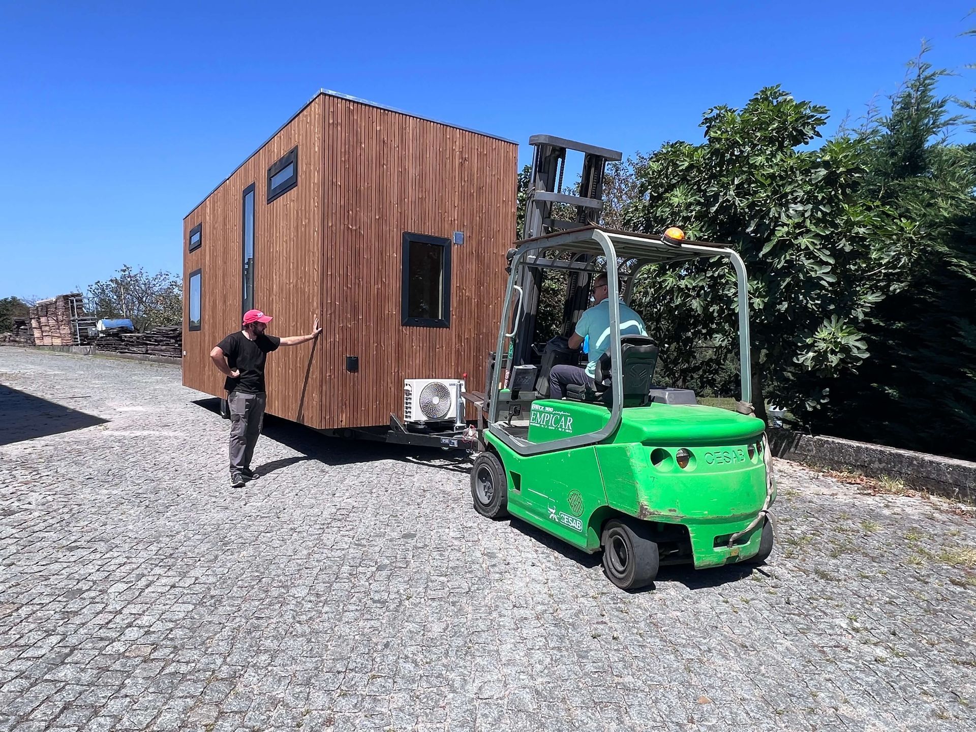 Forklift moving a small brown cabin on a gravel lot; a man in pink hat directs.