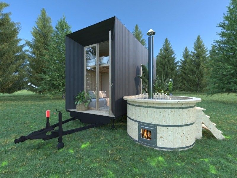 Tiny black cabin on a trailer with open door, next to a wood-fired hot tub outdoors.