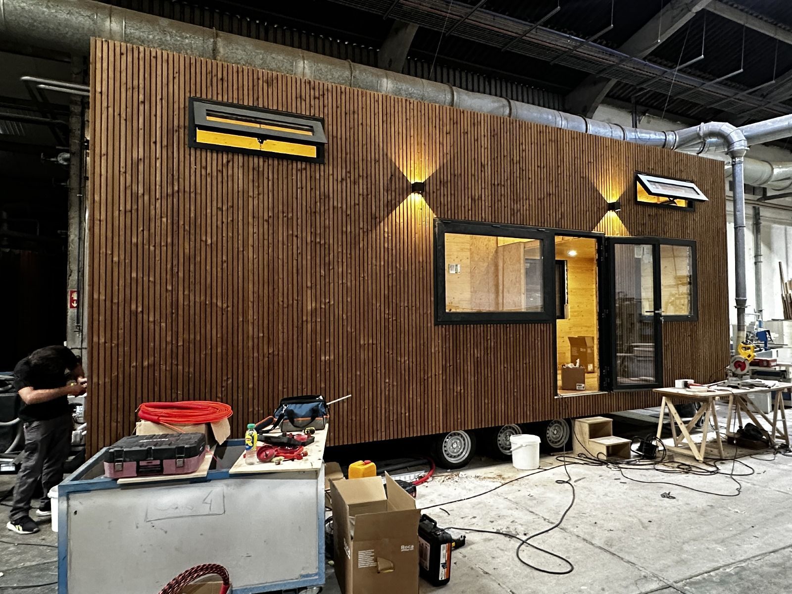 A wooden tiny house under construction inside a workshop. A person works on the outside.