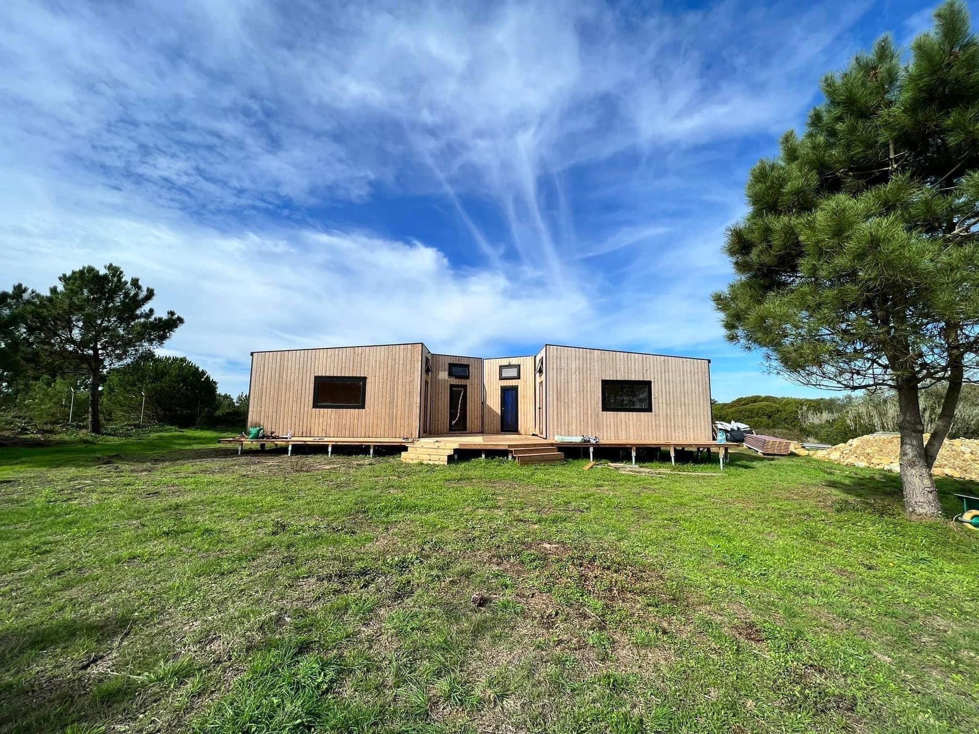 Wooden cabin under construction on a grassy hill, with blue sky and scattered clouds.