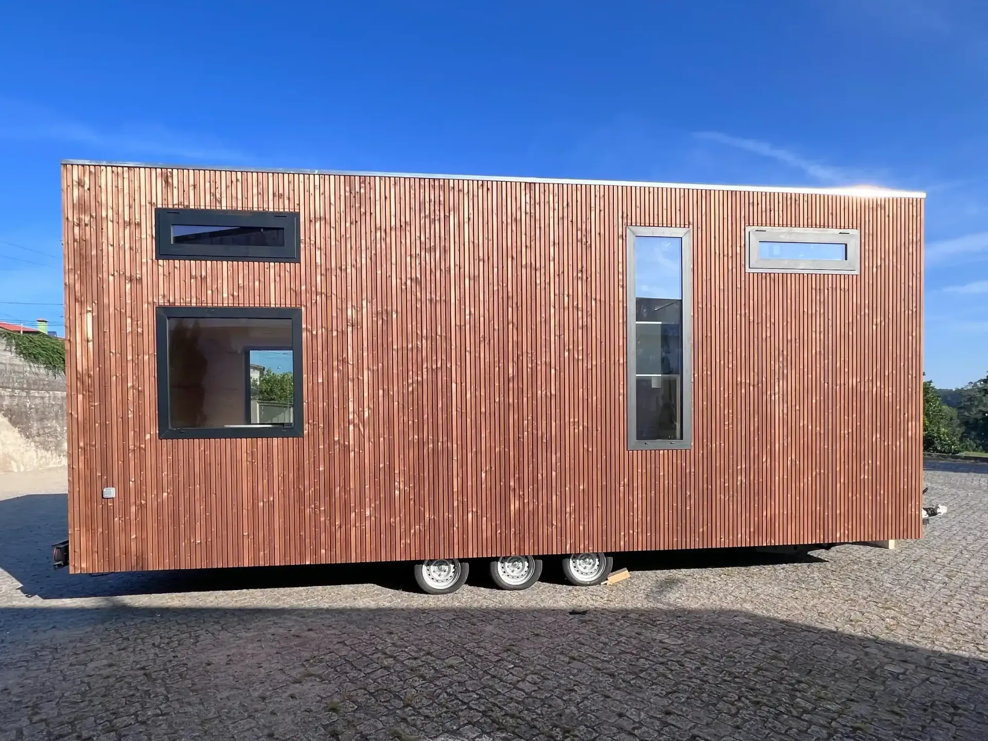 Wooden tiny house on a trailer with black-framed windows, against a blue sky.