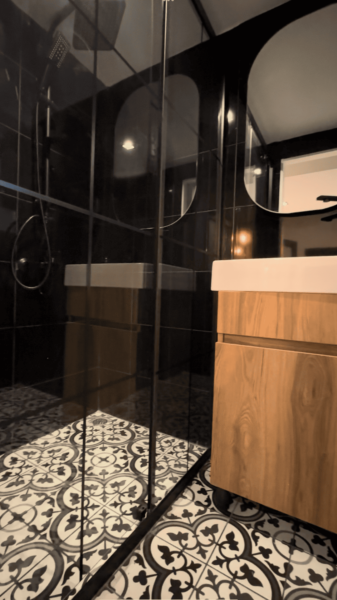 Black and white patterned bathroom with a glass shower door, wooden vanity, and black tiled walls.