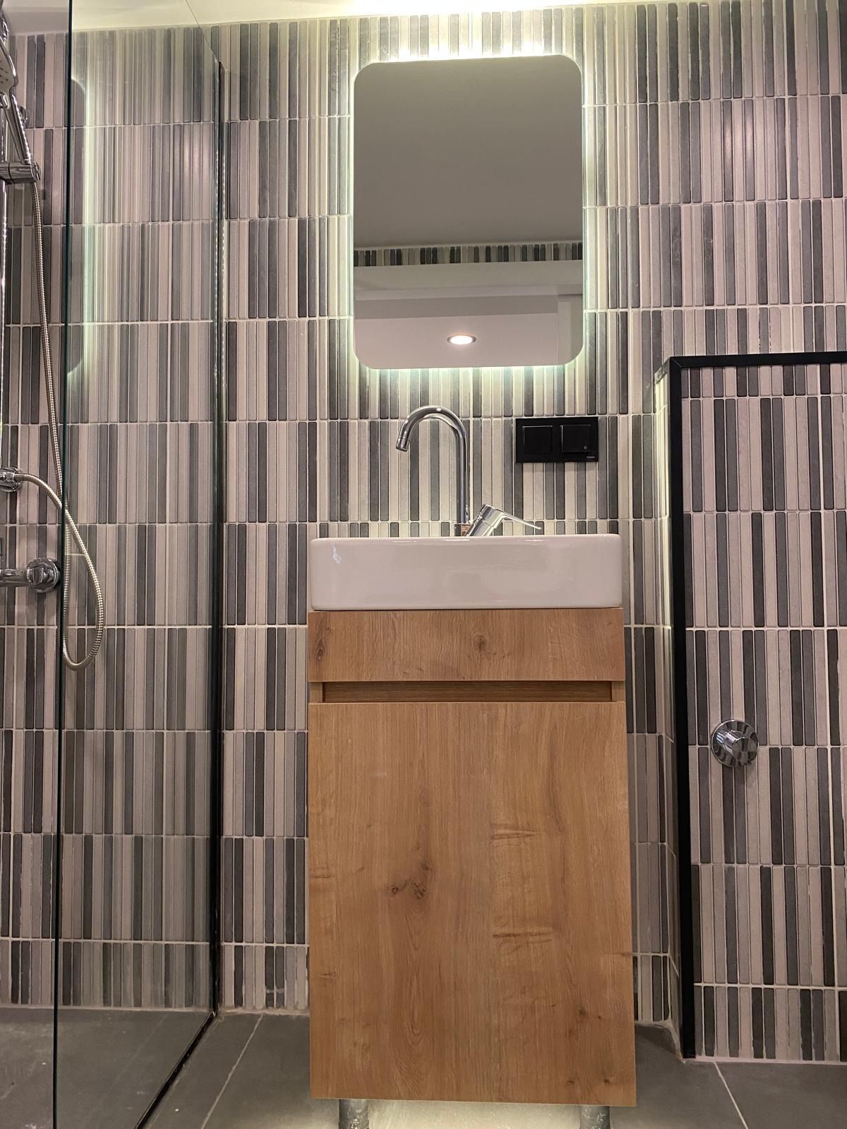 Bathroom with wood vanity, rectangular mirror with light, gray and white tile, shower on the left.