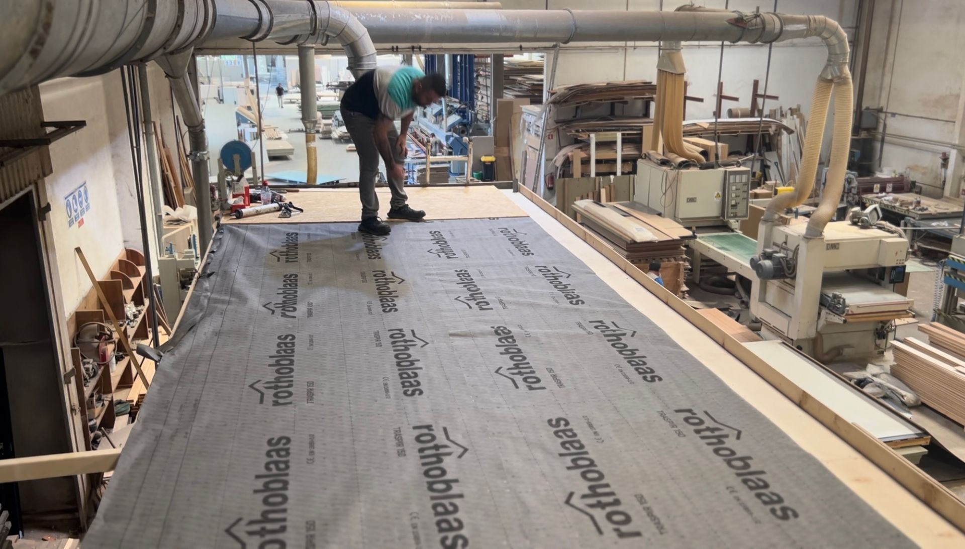 A person in a workshop installing dark membrane on a large wooden surface, other machinery in background.