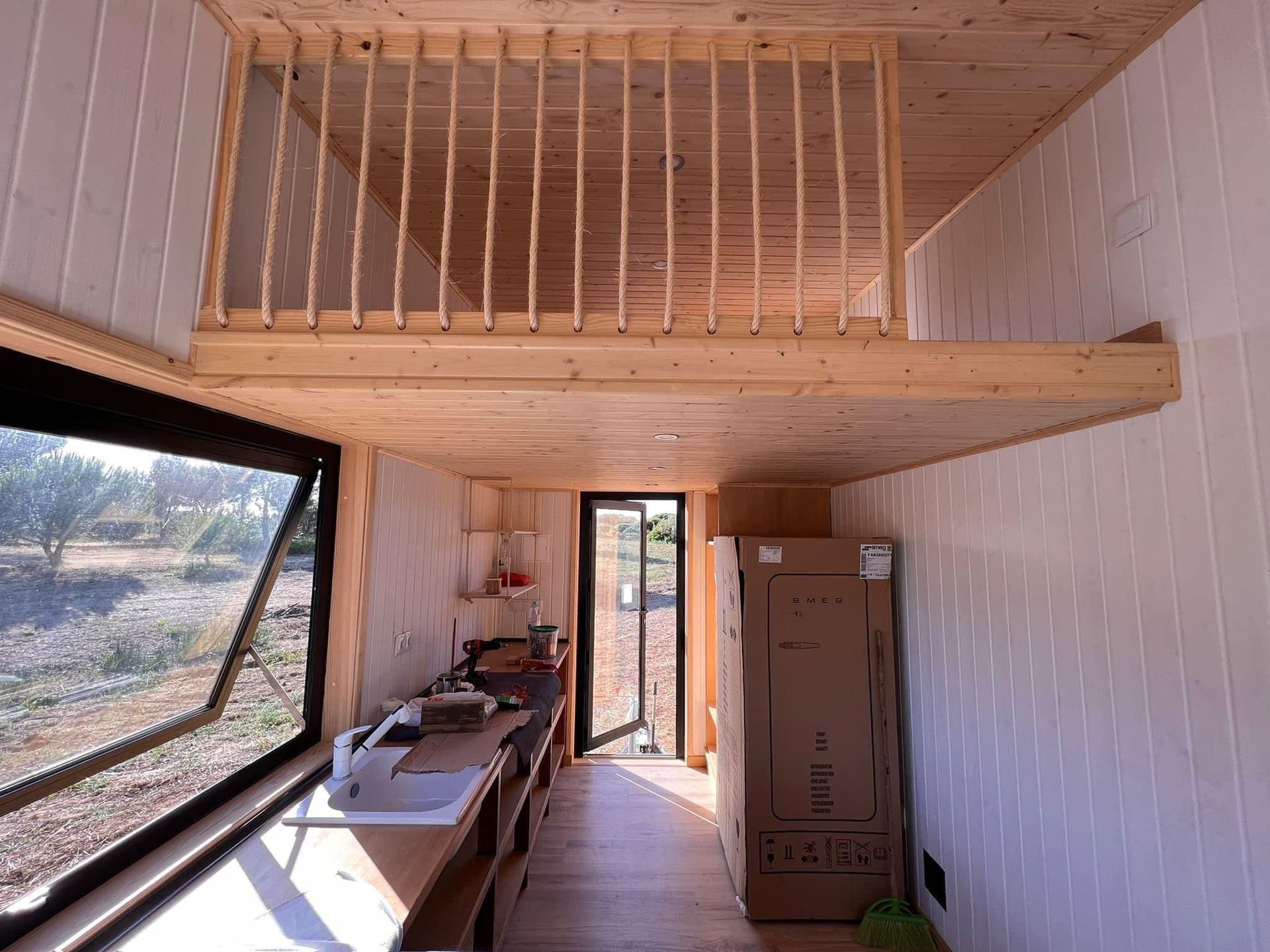 Interior of a tiny house with a loft, kitchen, and open window to the outside.