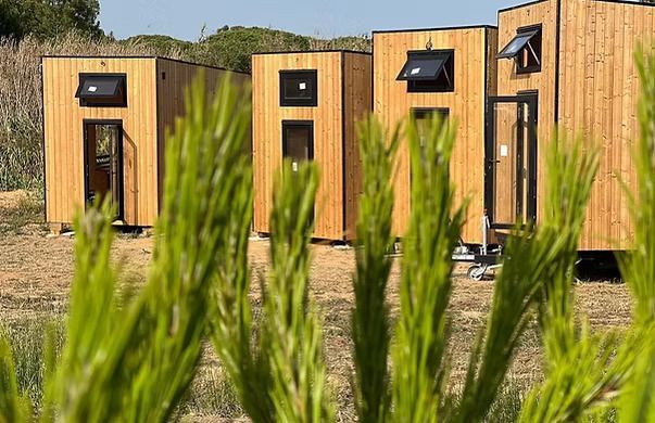 Wooden tiny houses, in a row, with dark frames, windows, and solar panels; set outdoors.