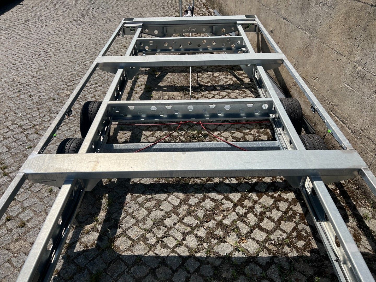 Metal trailer frame with four wheels, on cobblestone ground.