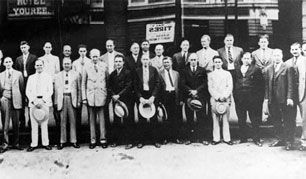Group of men outside a building, many holding hats. Building has a sign for Group of men outside a building, many holding hats. Building has a sign for