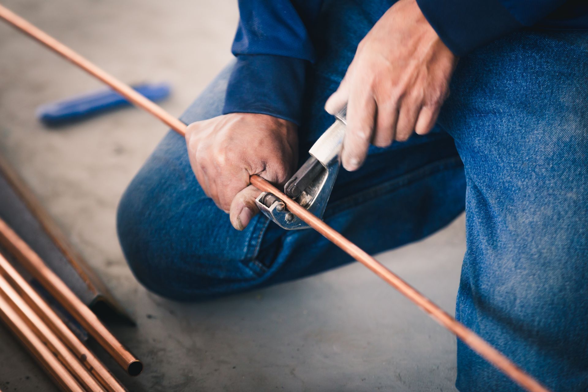 Person kneeling, cutting copper pipe with a pipe cutter. Blue jeans and shirt. Person kneeling, cutting copper pipe with a pipe cutter. Blue jeans and shirt.