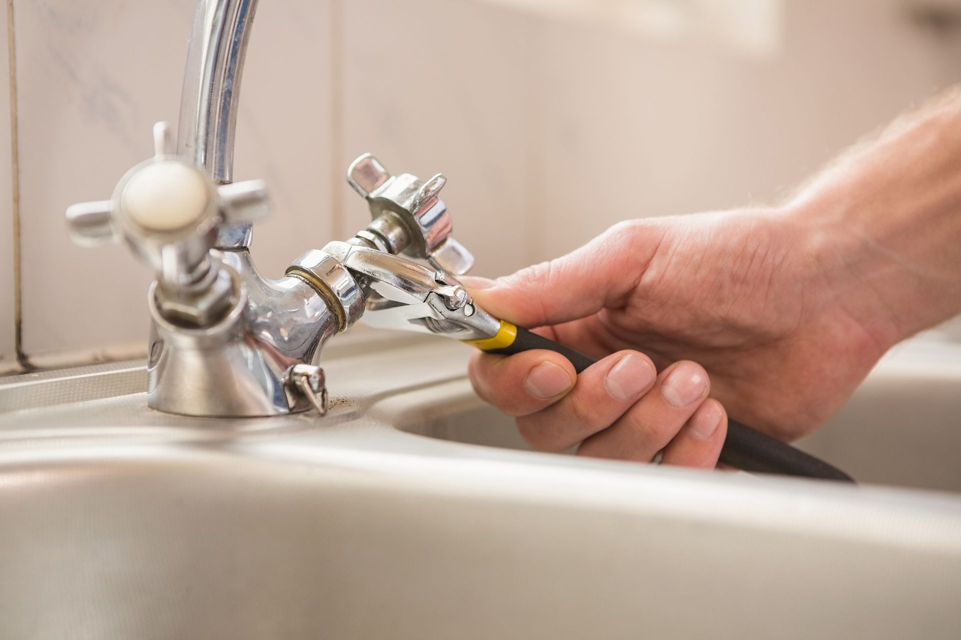 Person's hand using a wrench to fix a silver faucet in a stainless steel sink.