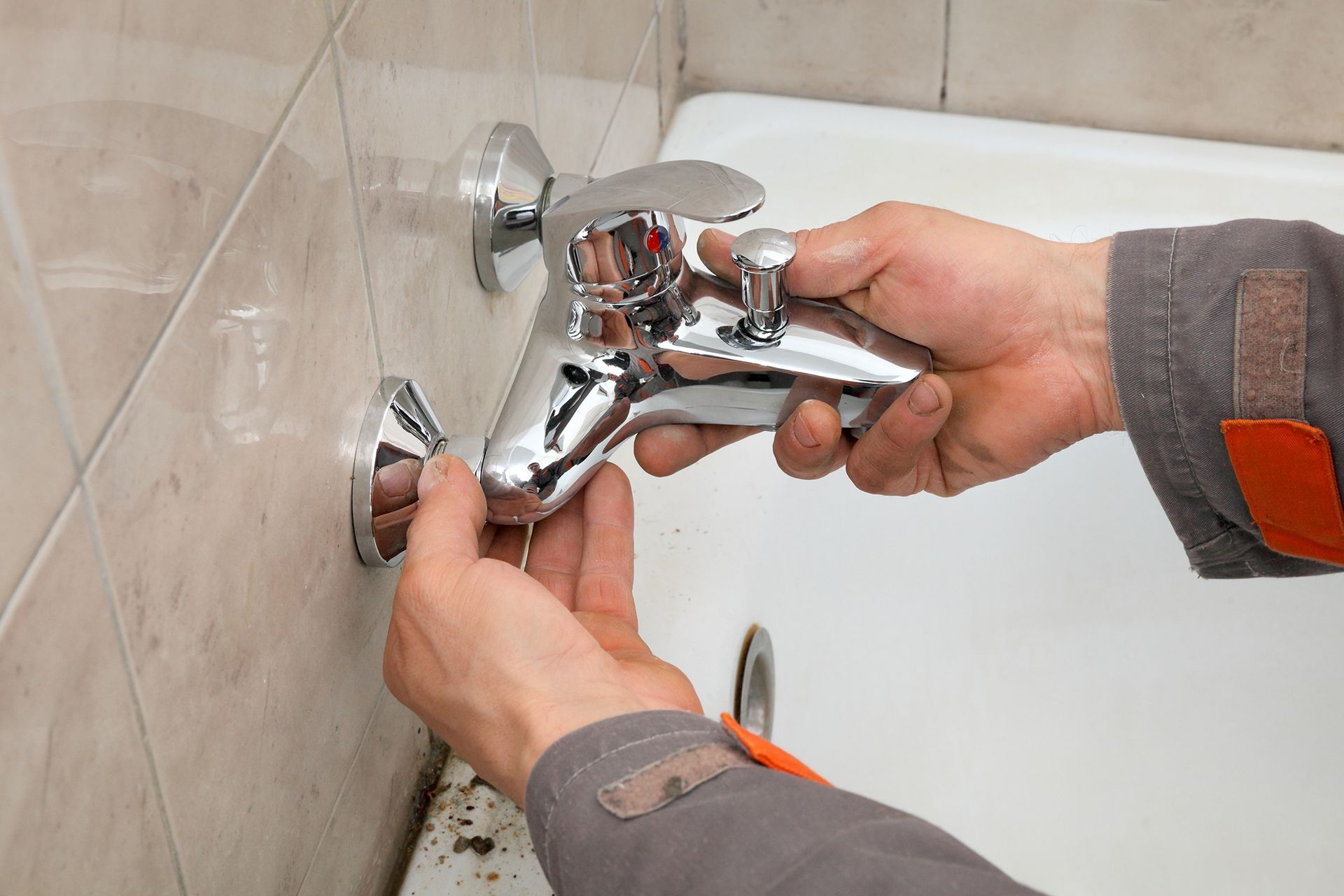 Plumber installing a chrome faucet on a tiled wall next to a bathtub.
