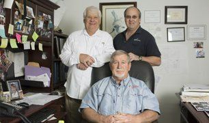 Three men in an office setting: one seated in a chair, two standing behind him, smiling. Three men in an office setting: one seated in a chair, two standing behind him, smiling.