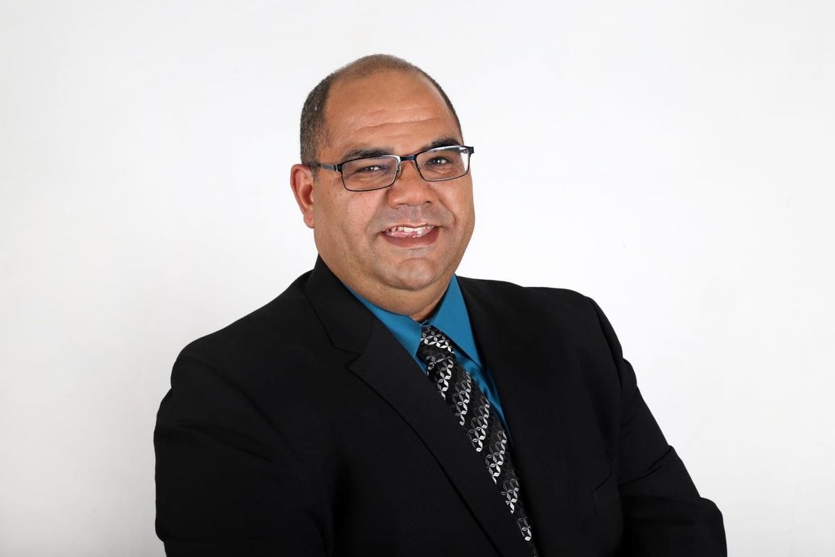 Smiling man in a black suit, blue shirt, and patterned tie against a white background