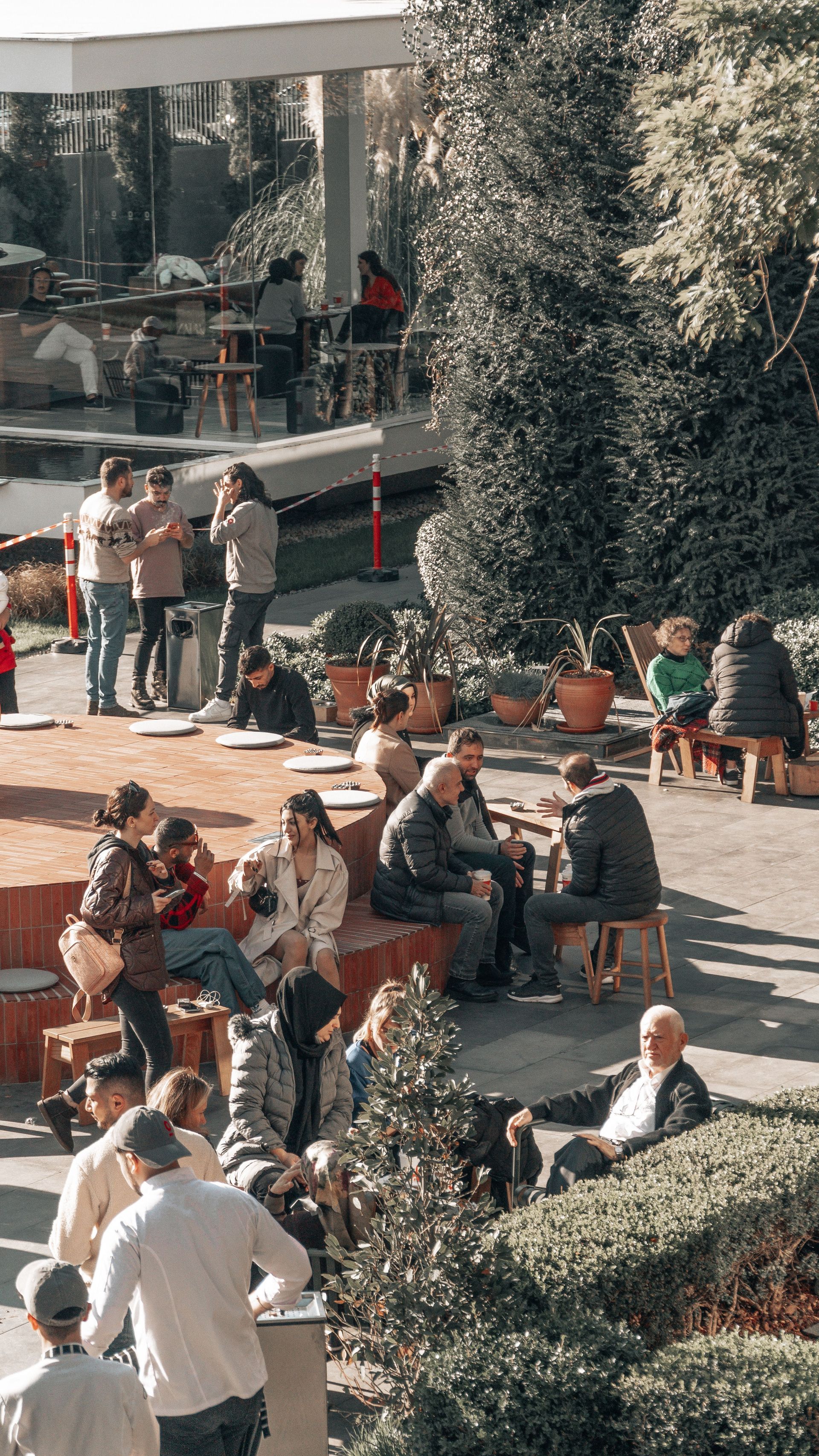 People sitting and standing in a sunny outdoor plaza with trees and benches.