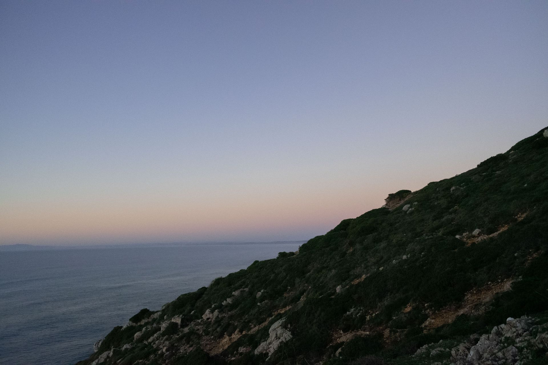 Dusk over a calm ocean with a dark, rocky hillside in the foreground