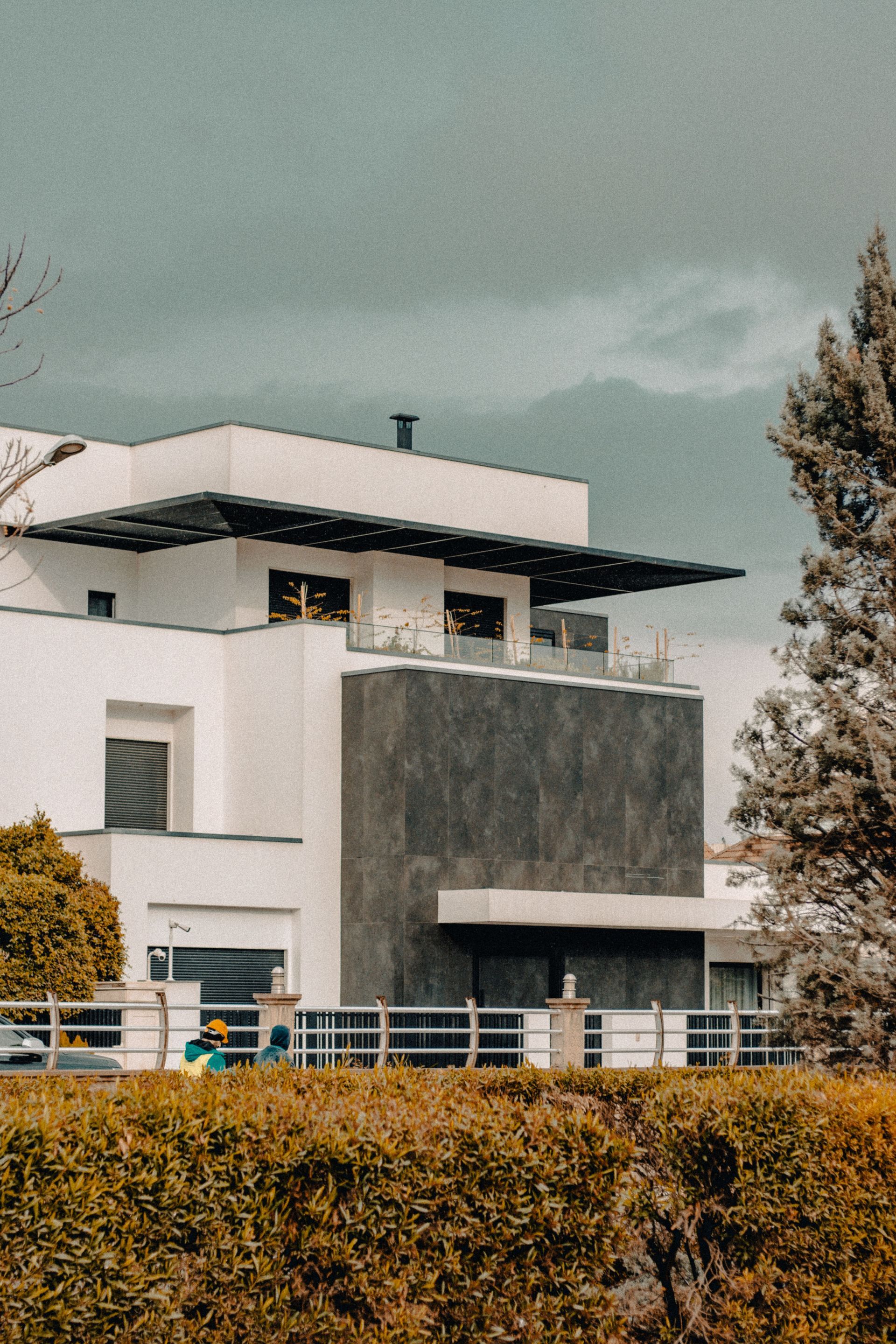 Modern white house with a dark gray section behind orange hedges under a cloudy sky