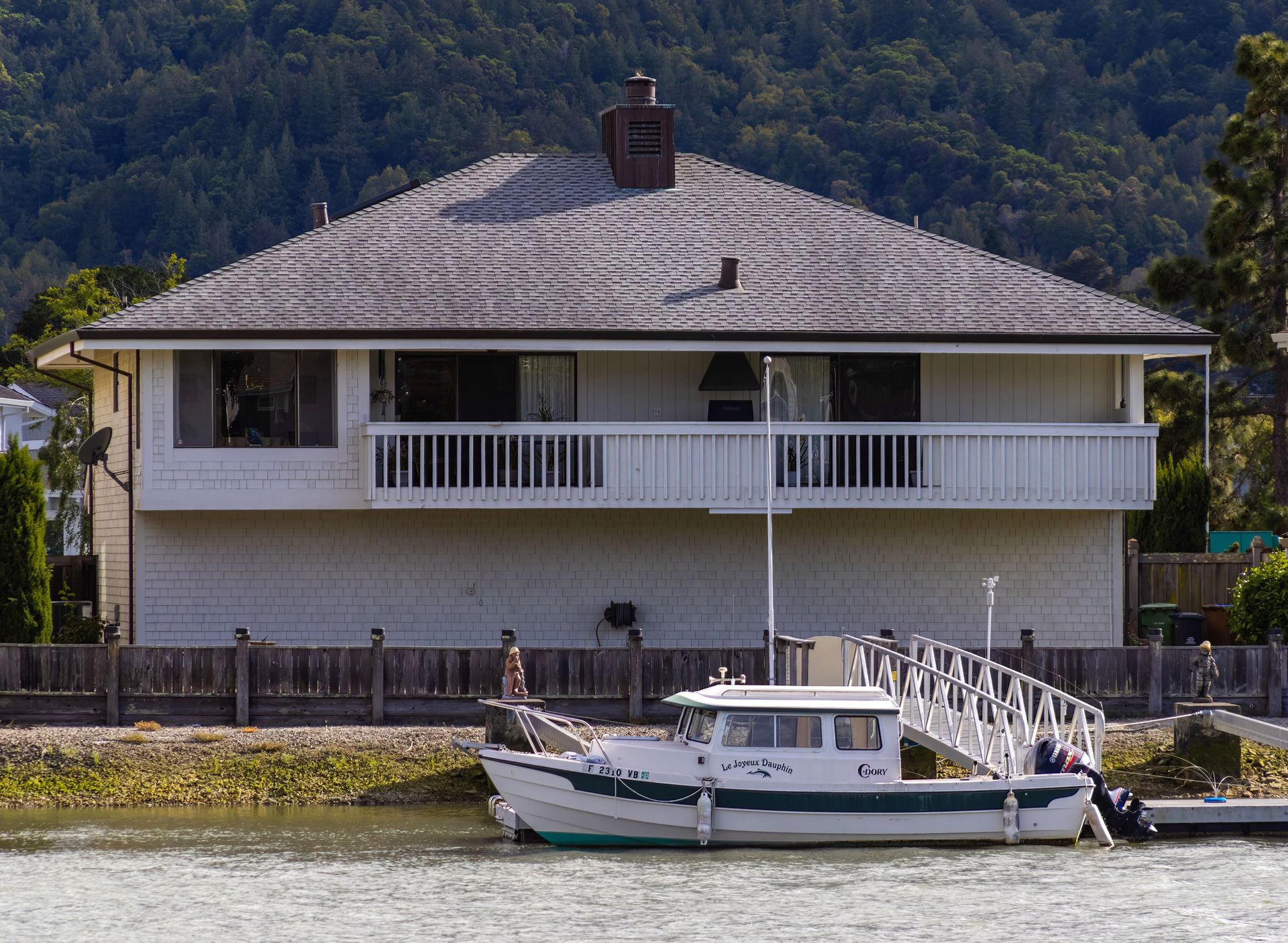 White waterfront house with balcony and a boat docked in front, backed by wooded hills
