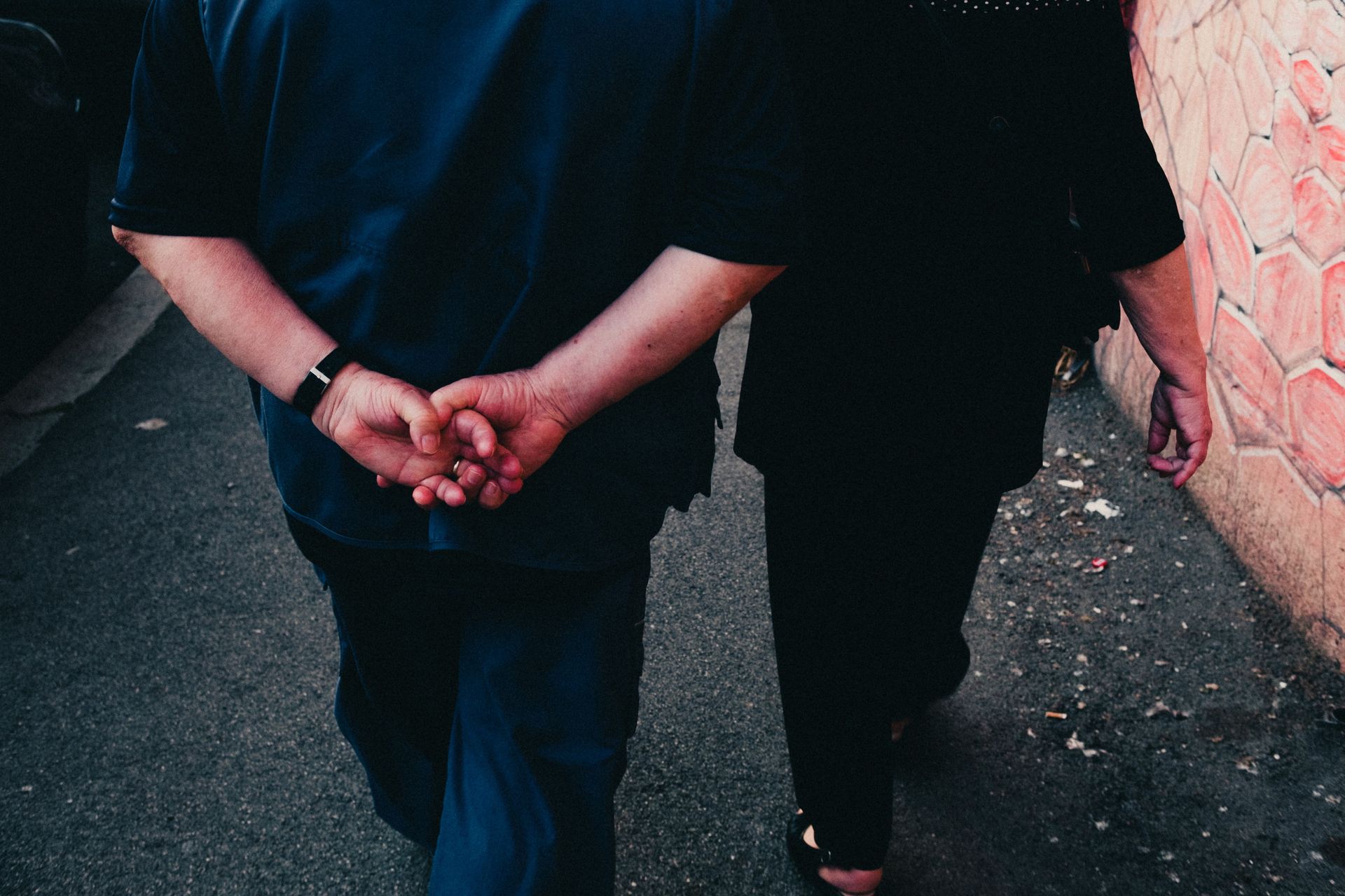 Two people walking hand in hand along a paved path beside a brick wall