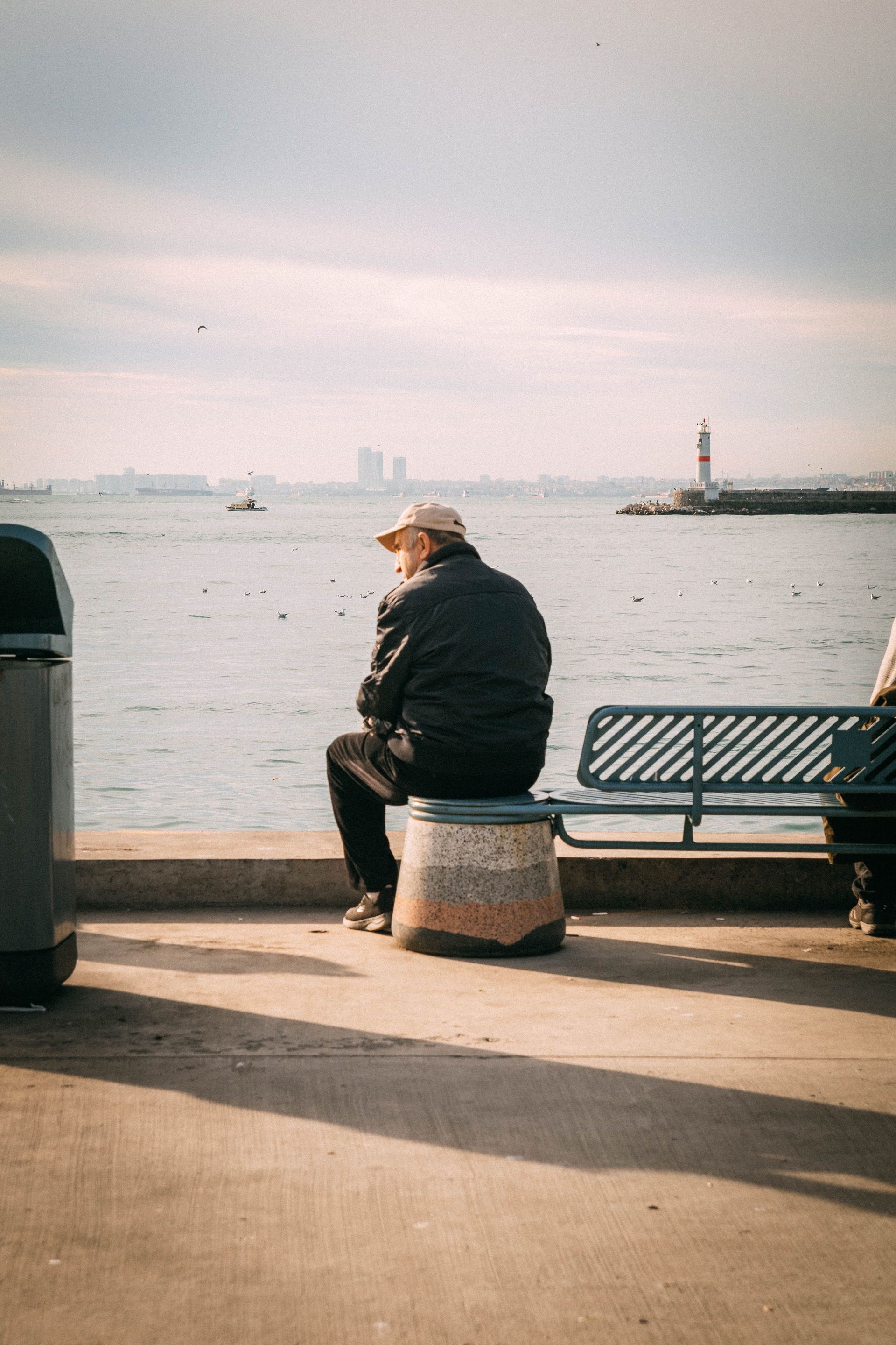 Person sitting on a bench by the waterfront, facing the water near a distant lighthouse.