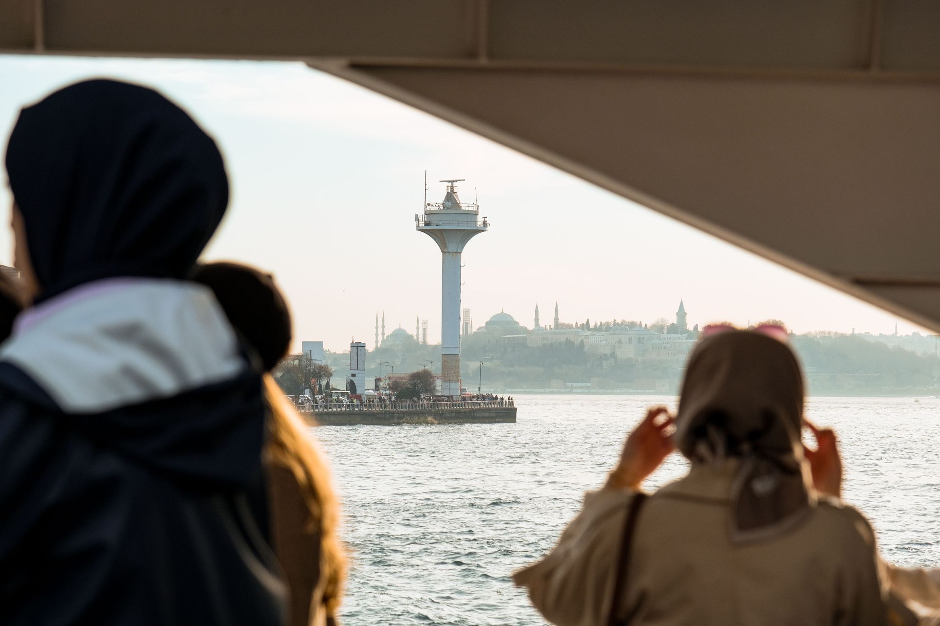 People on a ferry look out at a lighthouse and distant city skyline over the water.