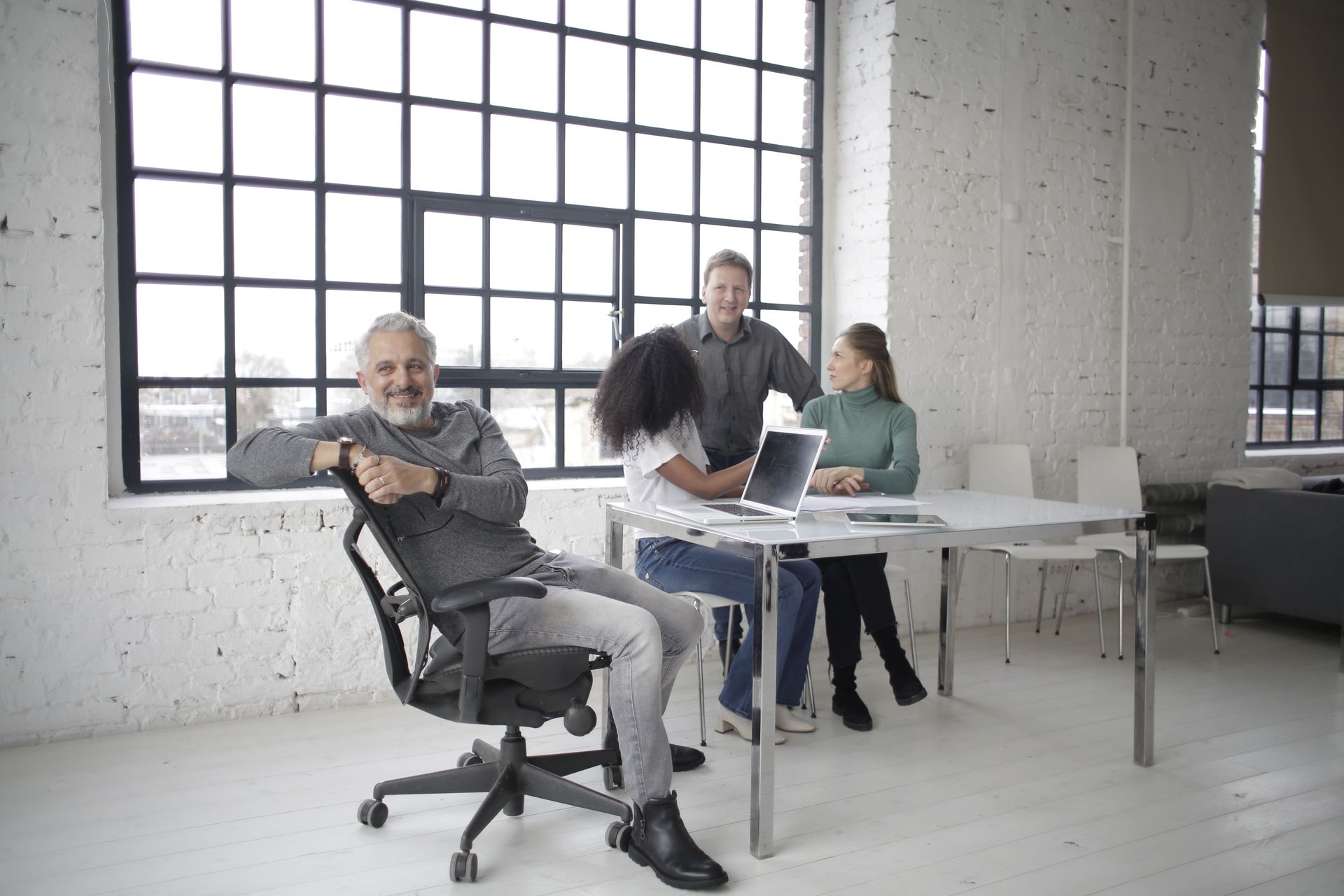 Four coworkers gathered around a white table in a bright loft-style office with large windows.
