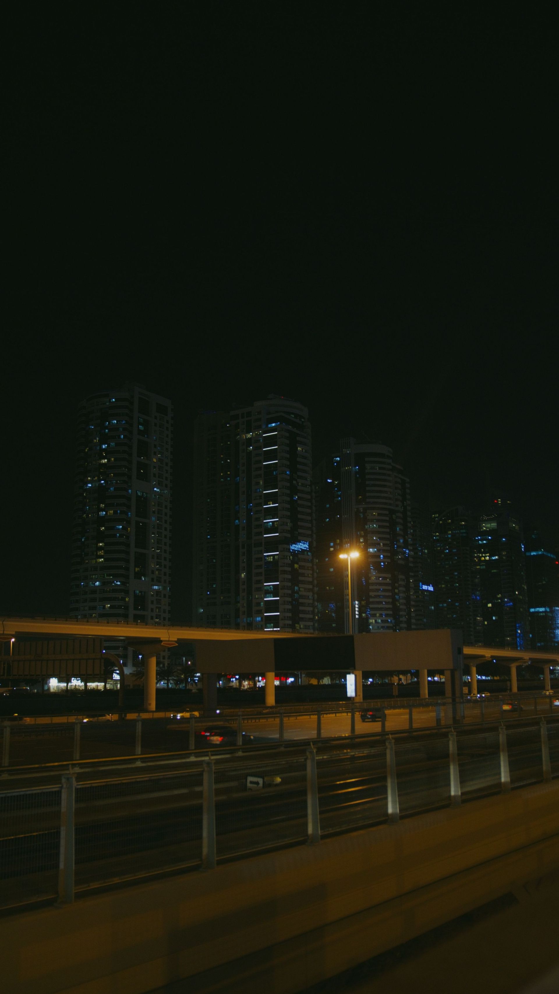 Night city skyline with lit high-rise buildings and a streetlight above a road and railing.