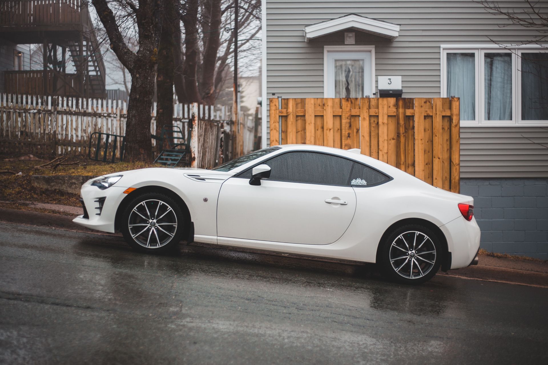 White sports car parked on a wet street beside a gray house and wooden fence