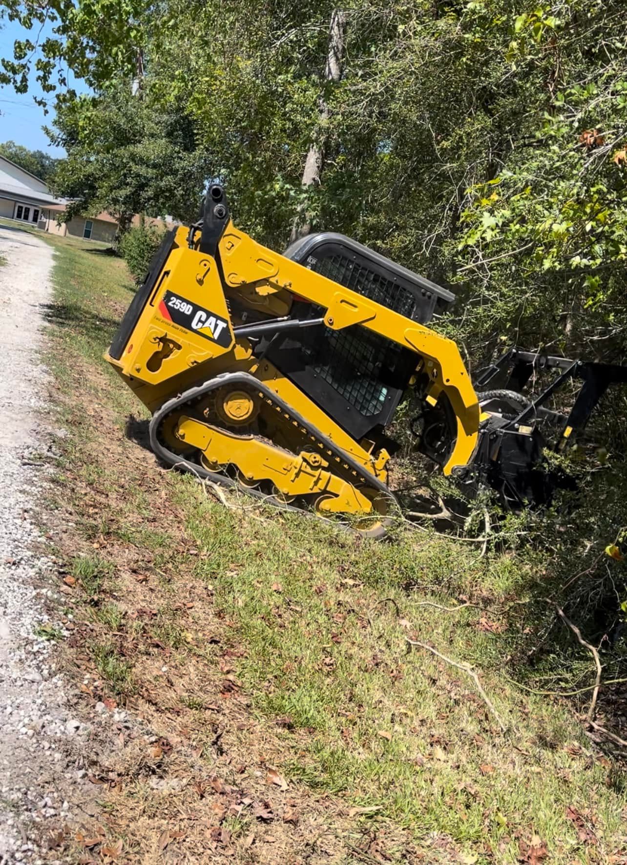 Yellow Caterpillar skid steer angled on a grassy slope next to trees and a road.