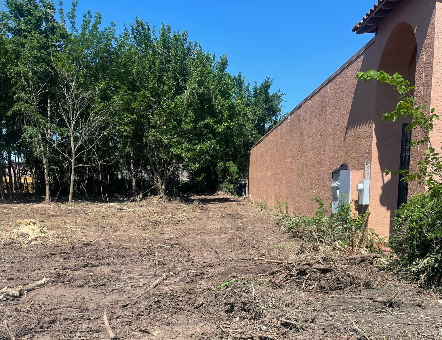Cleared dirt area next to a building with trees in the background under a blue sky.