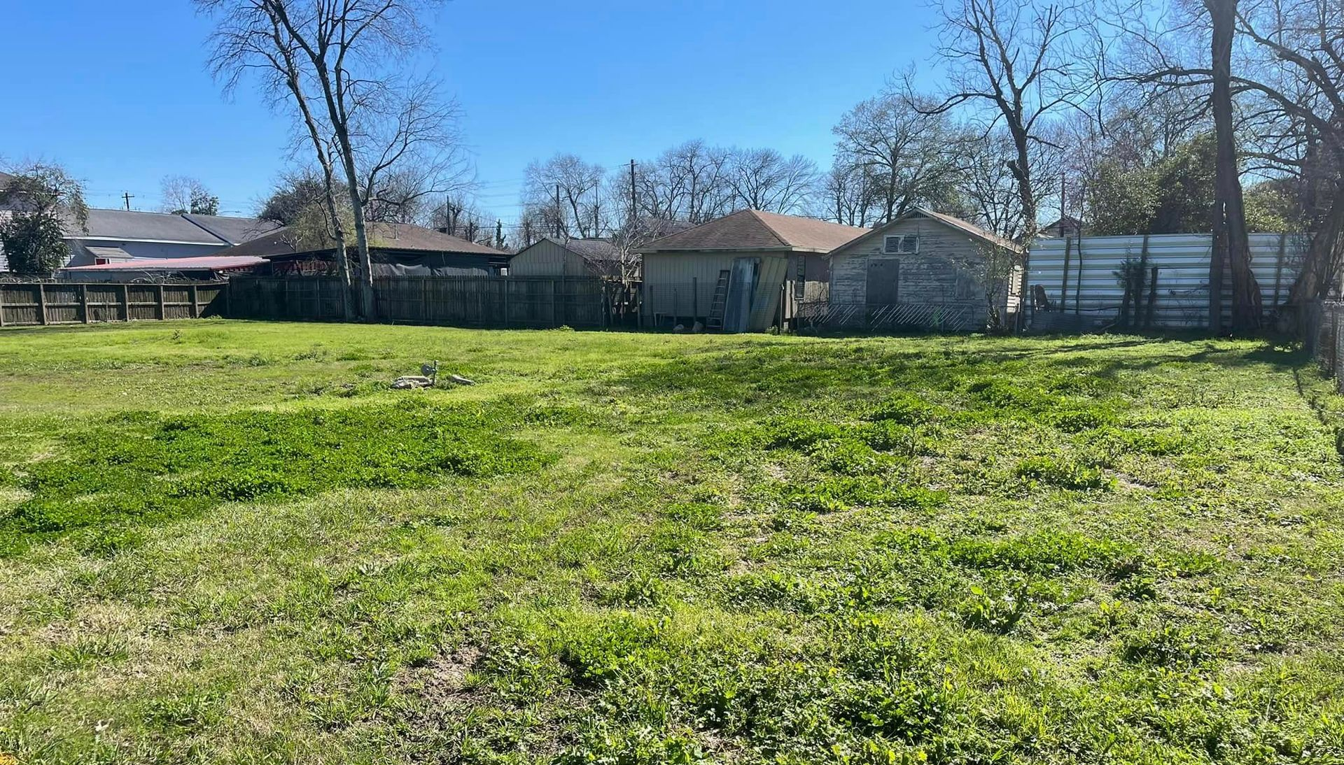 Grassy vacant lot with small structures and fence in the background under a blue sky.