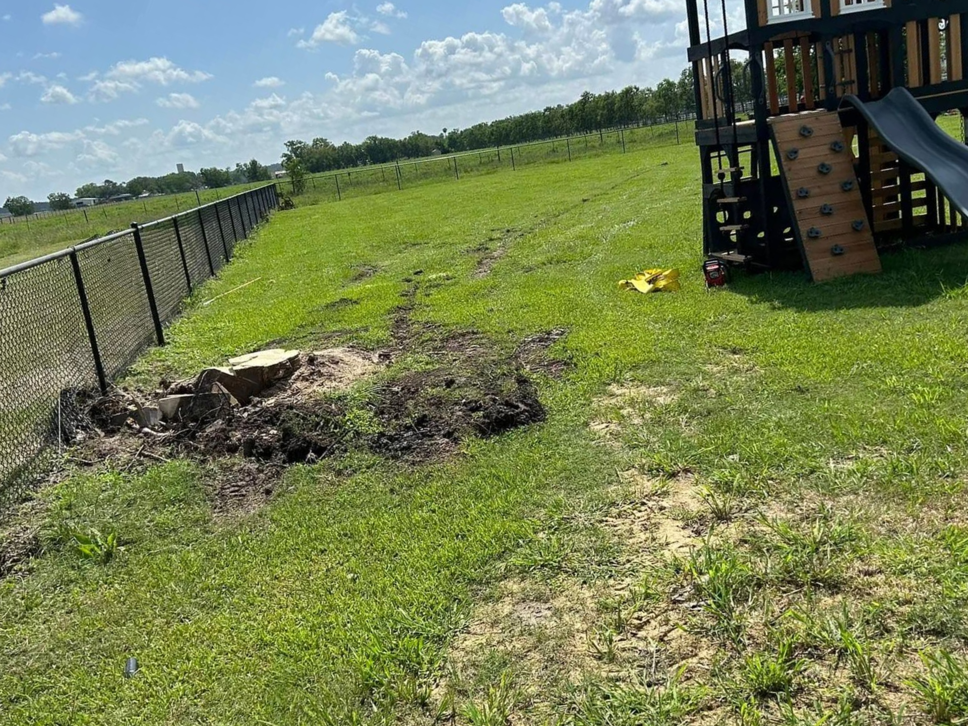 Backyard with a chain-link fence, green grass, a playground, and disturbed soil.