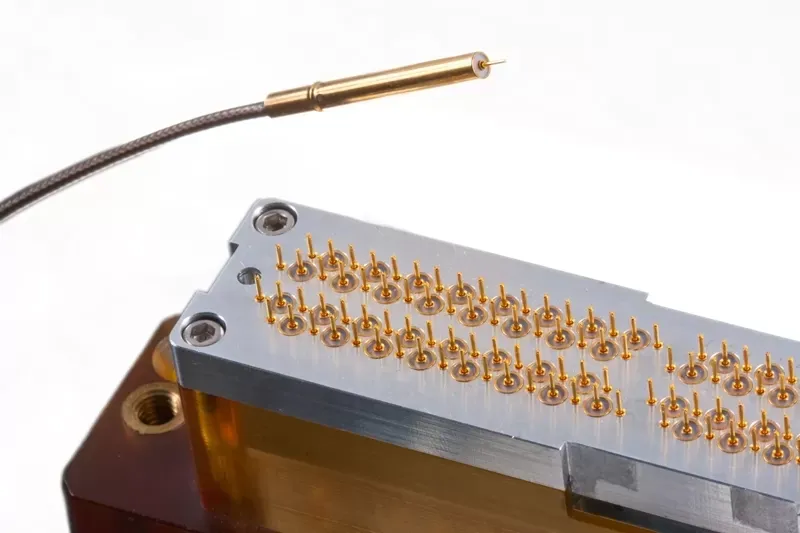 A gold probe needle cable rests above a silver test socket block containing dozens of vertical gold spring-loaded pins.