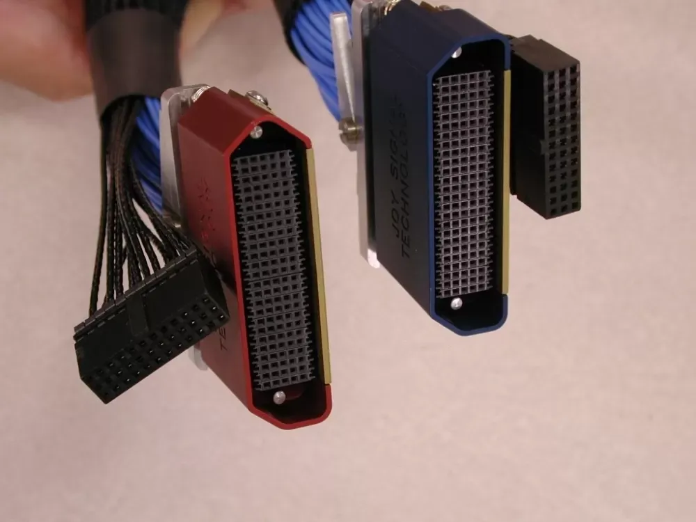 Two specialized, multi-pin electrical connectors, one red and one blue, held by hand with attached black ribbon cables.
