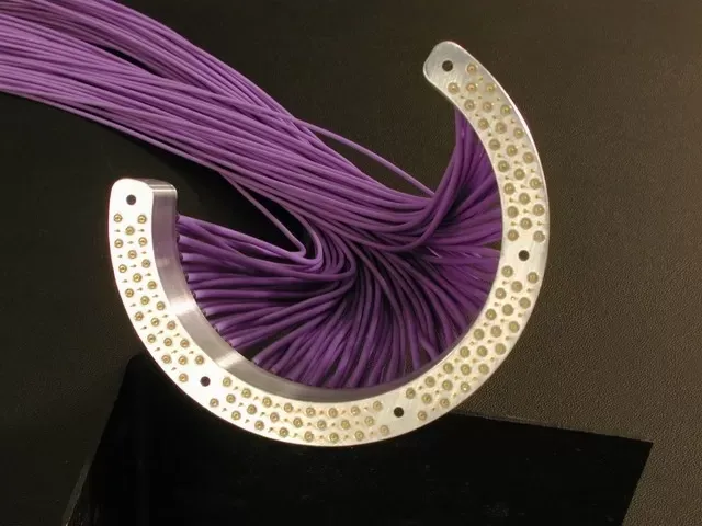 A metallic, crescent-shaped device with many holes holds a dense bundle of purple fiber optic cables.