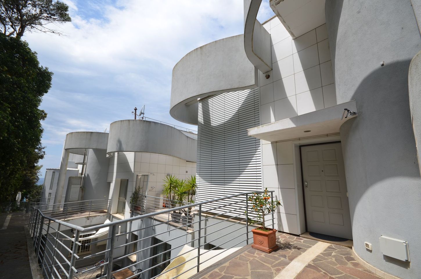 Terrazza di un moderno edificio bianco con pareti curve, ringhiere in metallo, scale e piante in vaso sotto un cielo azzurro.