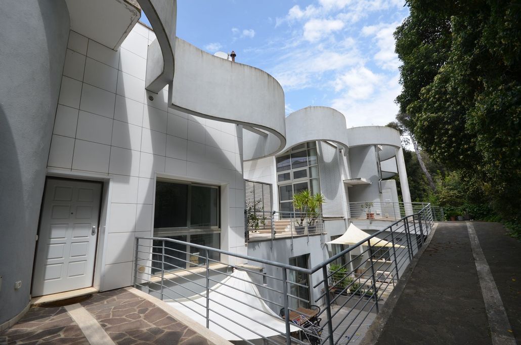 Moderno edificio bianco con pareti curve e passerelle lungo un sentiero alberato sotto un cielo parzialmente nuvoloso.