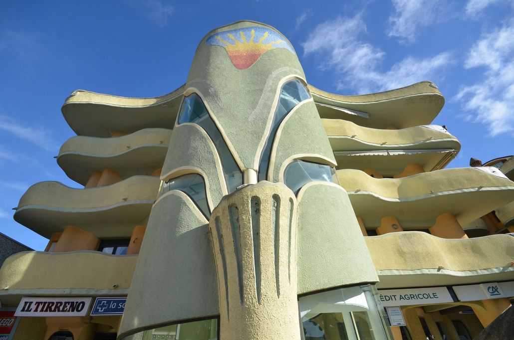 Facciata colorata in stile art déco con balconi curvi e una torre centrale scolpita che si staglia contro un cielo azzurro.