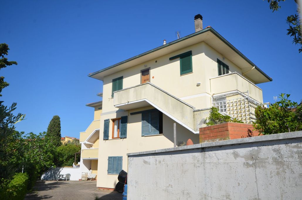 Casa a due piani color crema con persiane verdi, scale esterne e una parete bianca sotto un cielo azzurro.