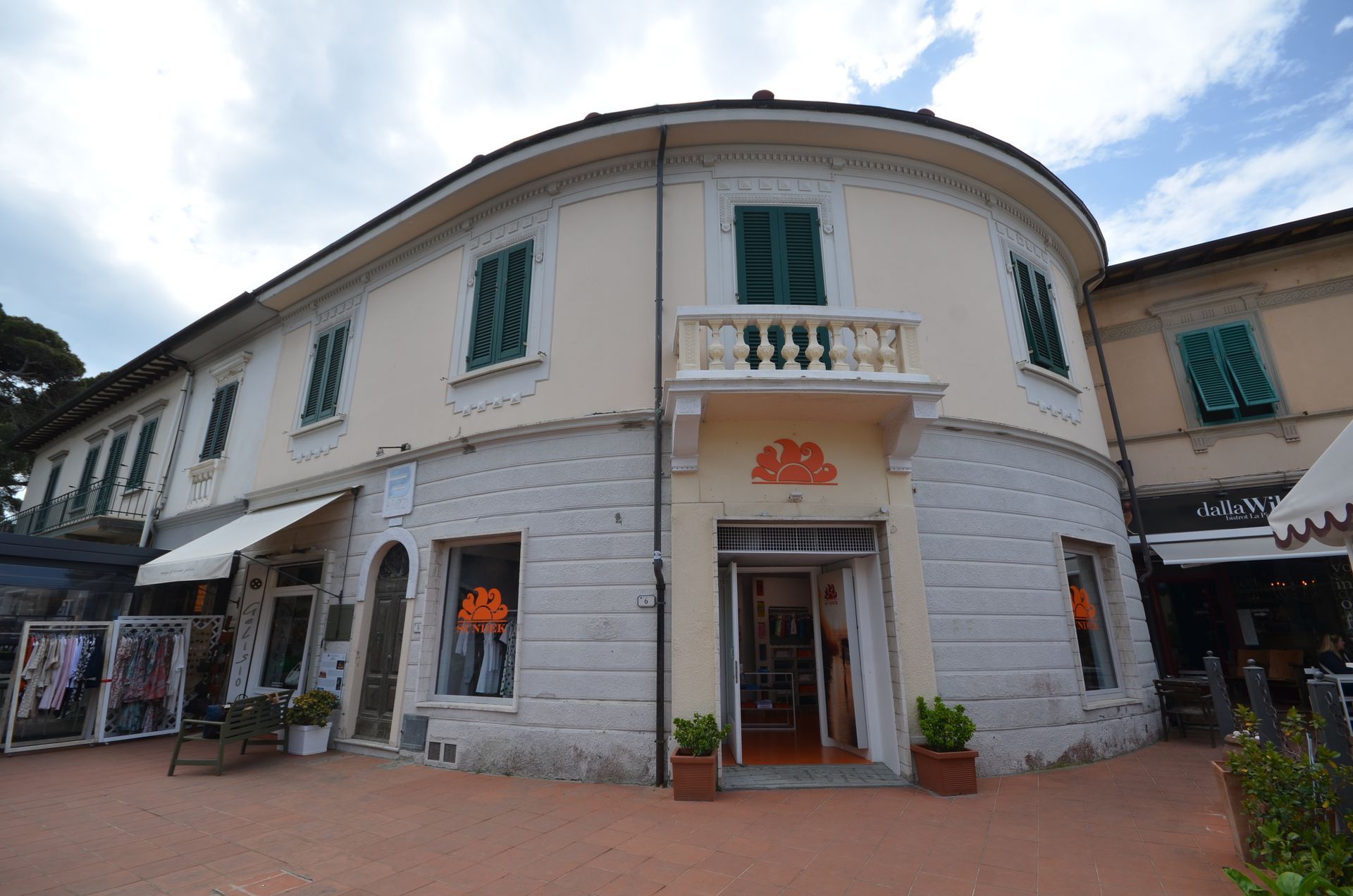 Edificio bianco curvo con persiane verdi e vetrine lungo un vialetto di mattoni sotto un cielo parzialmente nuvoloso.