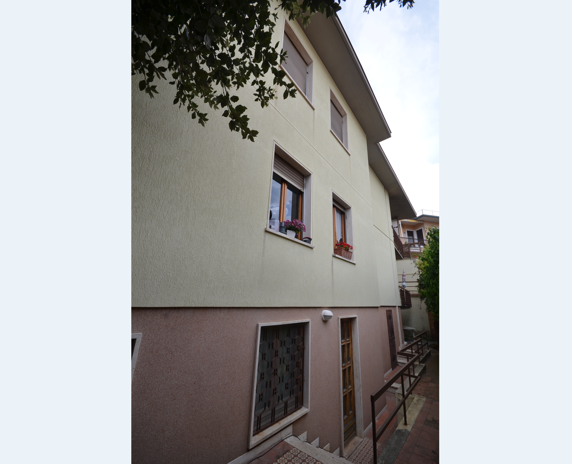 Vista laterale ravvicinata di una casa in stucco beige con finestre incorniciate di rosso e un vialetto in pendenza accanto ad essa.
