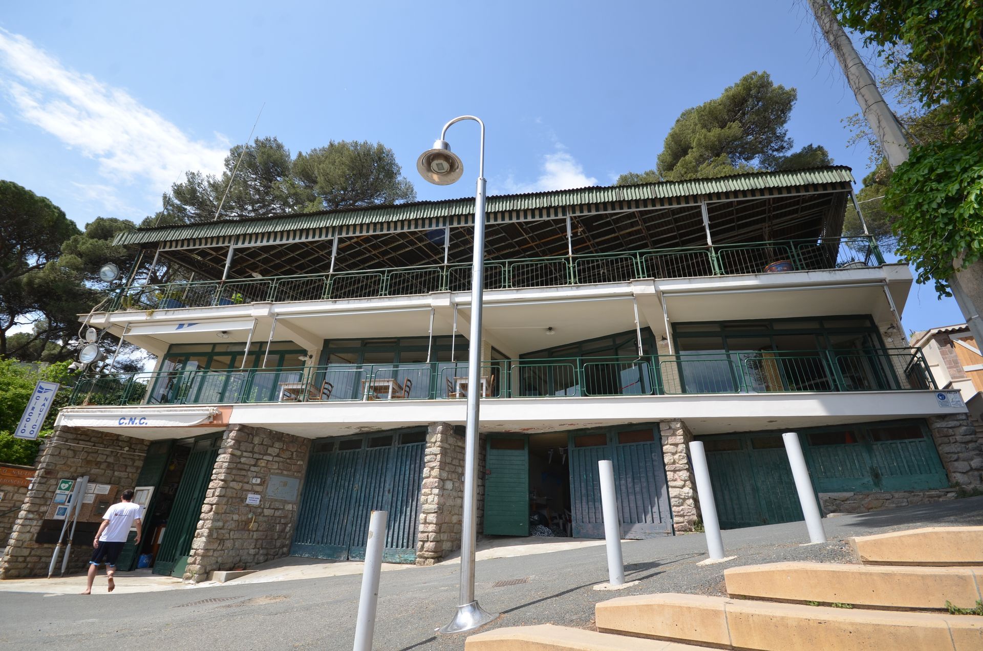 Esterno di un edificio moderno con muri in pietra, balconi in vetro e un tetto con pannelli solari sotto un cielo azzurro.