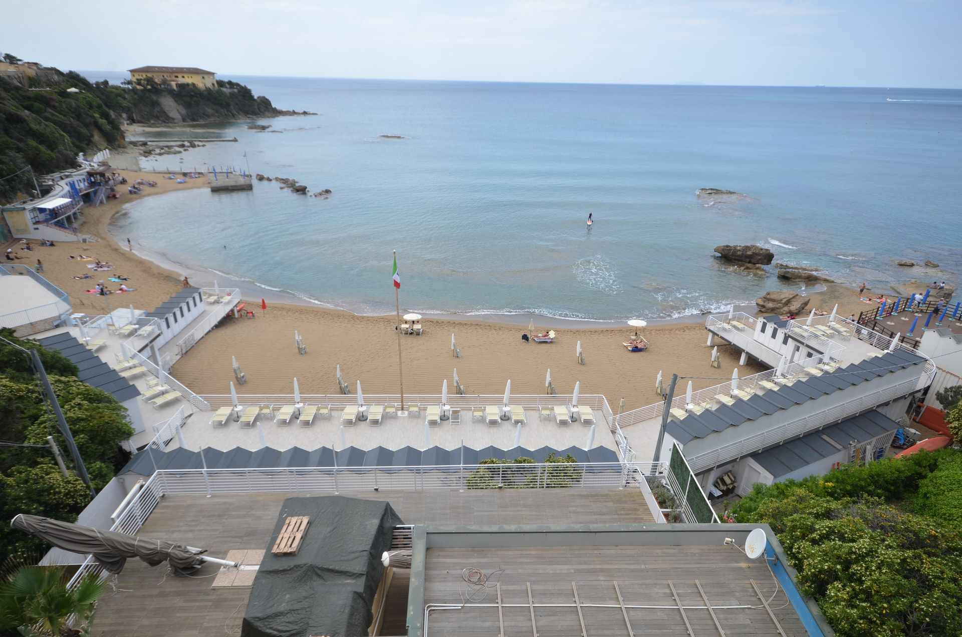 Vista dall'alto di un resort balneare con una baia sabbiosa a forma di mezzaluna, mare calmo e lettini e ombrelloni bianchi.